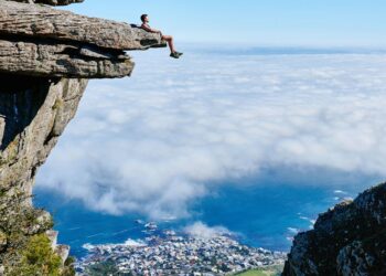 person sitting on mountain cliff: The poem opens with the personification of nature, framing climate change as a message—an alarm that humanity must heed. This sets the tone: the Earth is in crisis, and it is calling out for rescue.